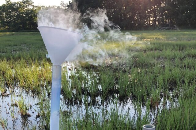 A funnel in a pipe releases steam against the backdrop of a flooded salt marsh.