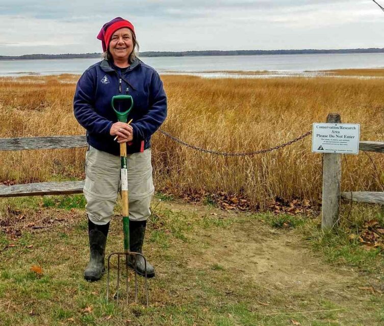 A woman in a red hat with a pitchfork standing in front of a salt marsh conservation research area.