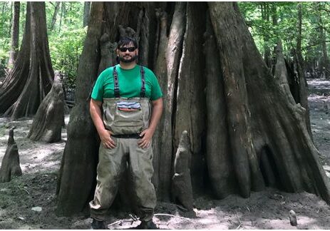 A man in waders and sunglasses posing in front of a tree three times as wide as him.