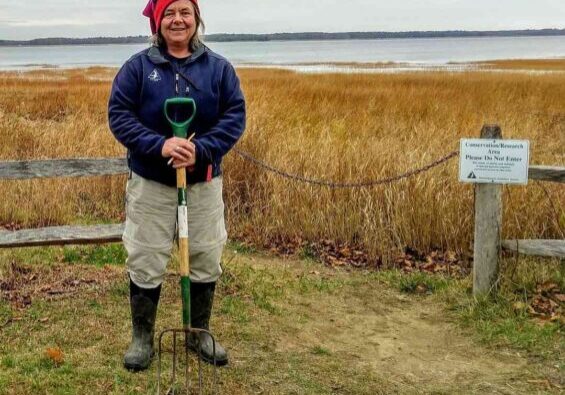 A woman in a red hat with a pitchfork standing in front of a salt marsh conservation research area.