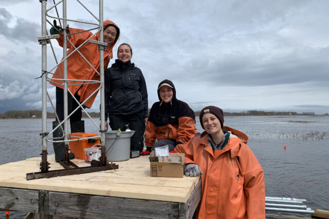 Four scientists in black and orange jackets pose on and around a wooden platform with a metal tower.
